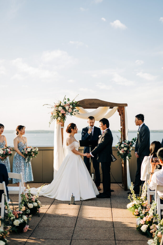 Bride and groom holding hands during lakeside ceremony at Edgewater wedding in Madison with floral arch and water backdrop.