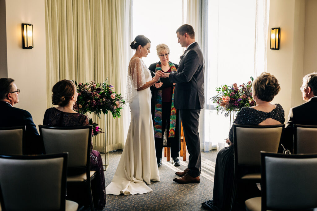 Couple exchanging rings during intimate indoor ceremony at Edgewater wedding in Madison with guests seated nearby.