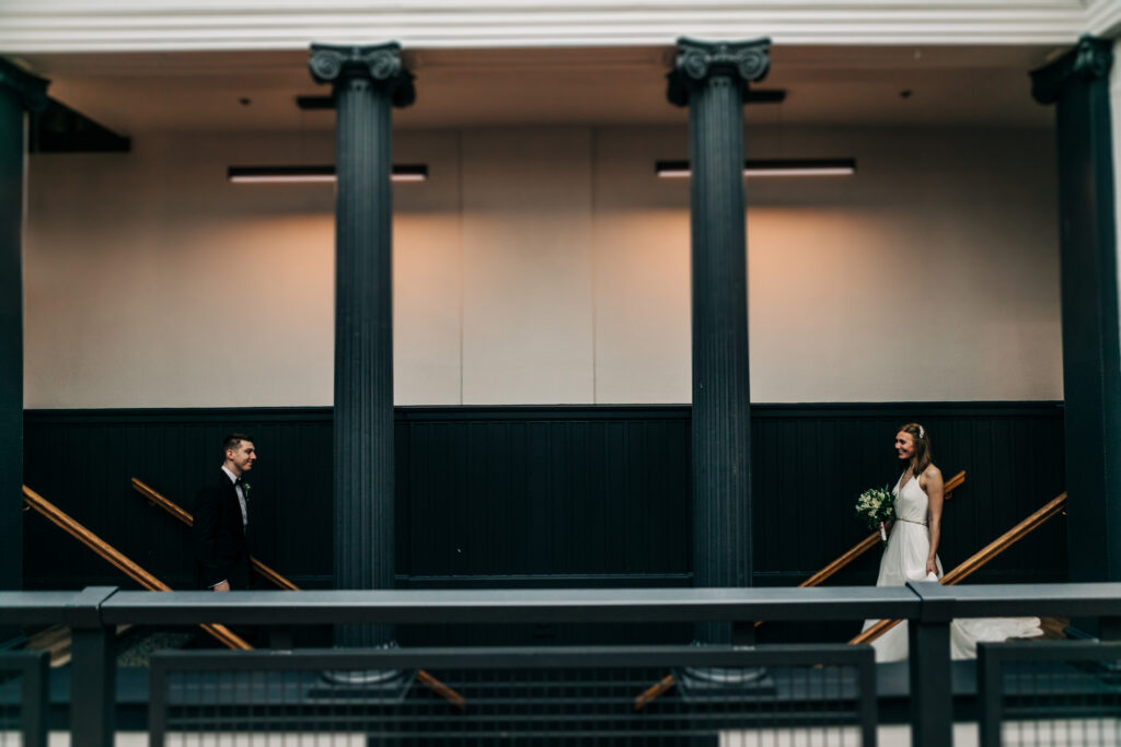 Bride and groom standing apart across balcony framed by tall columns inside Ellsworth Block wedding venue in Madison.