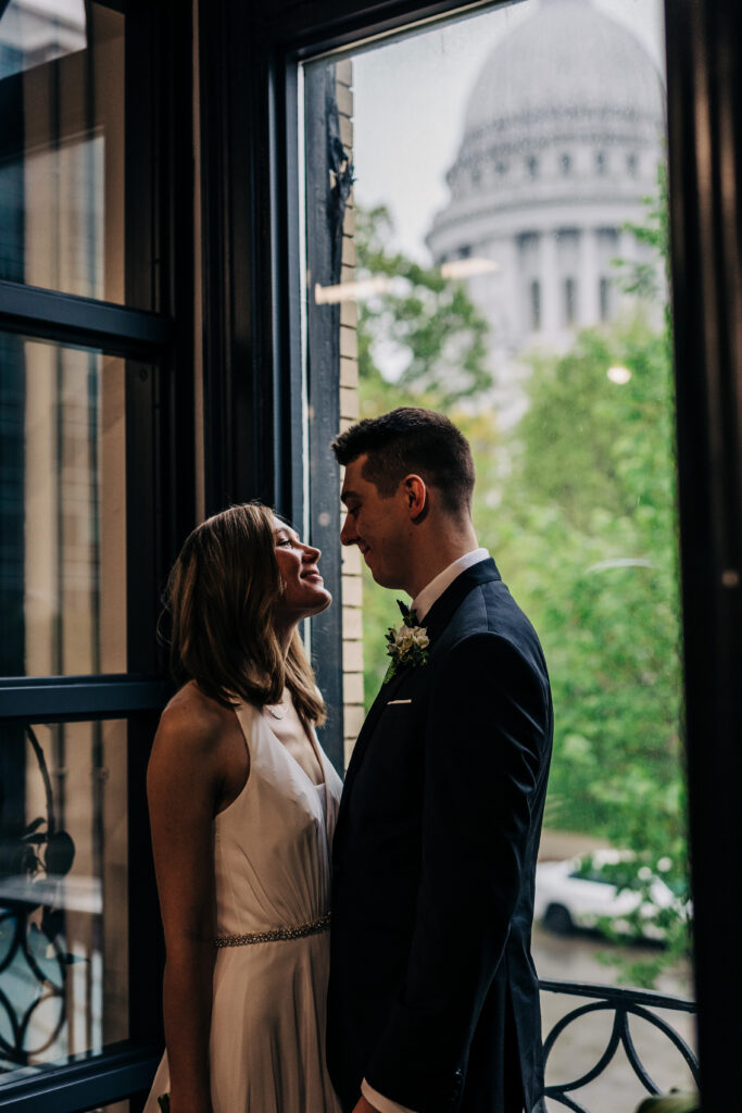 Bride and groom standing close by window with Wisconsin State Capitol in background at Ellsworth Block wedding in Madison.
