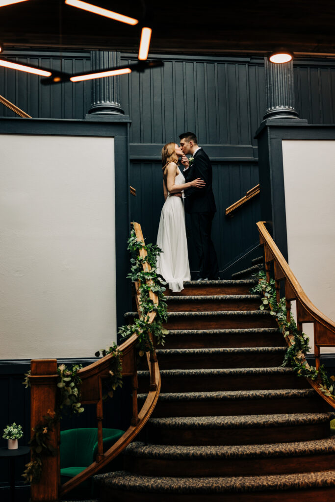 Bride and groom kissing on curved staircase decorated with greenery at Ellsworth Block wedding venue in Madison.
