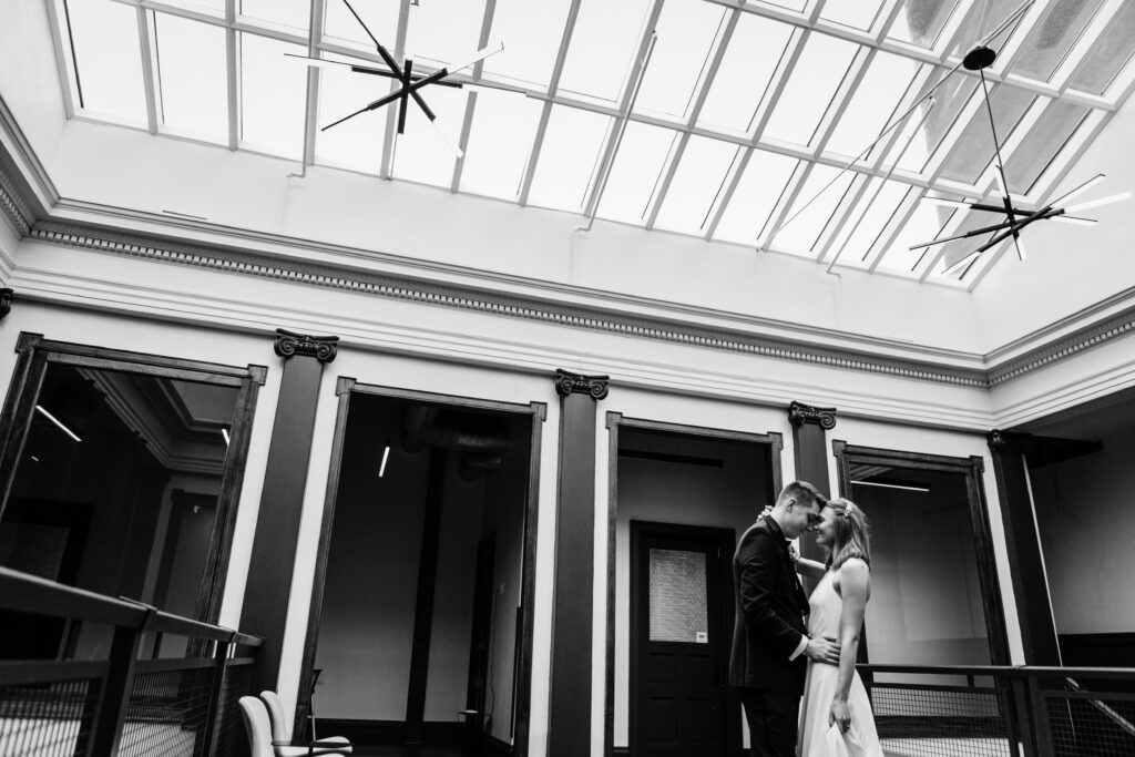 Bride and groom embracing on indoor balcony beneath glass ceiling at Ellsworth Block wedding venue in Madison, black and white portrait.