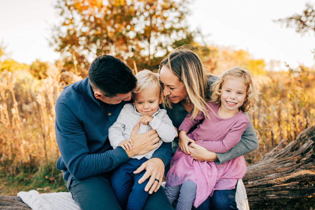 Family of four sitting on a log outdoors, parents holding children with autumn trees during Madison fall mini session.