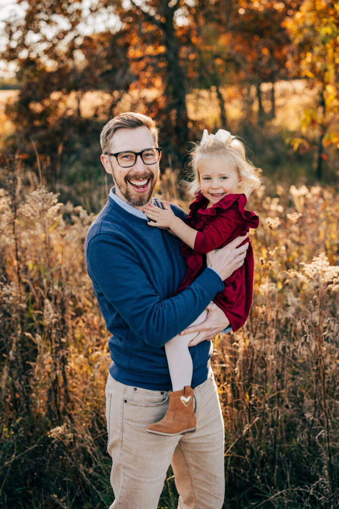 Father holding young daughter in a field with golden fall foliage during outdoor family photos in Madison WI.