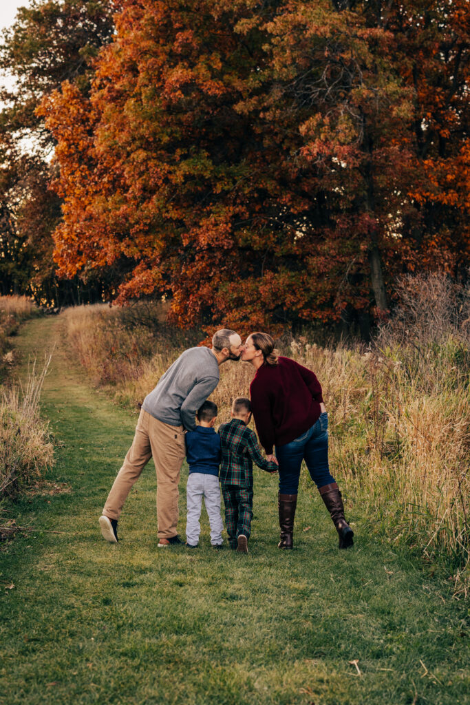 Parents walking on a path with two children and sharing a kiss under vibrant fall trees in Madison, Wisconsin.