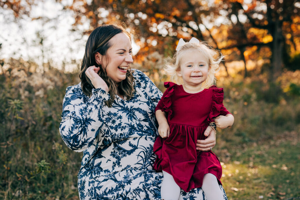 Mother sitting with toddler daughter in red dress, both smiling in a sunlit autumn field in Madison Wisconsin.