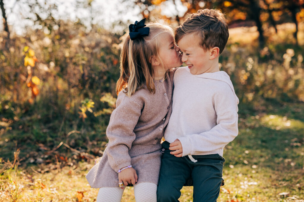 Young girl kissing a boy on the cheek while sitting together in a fall field during Madison WI family photos.