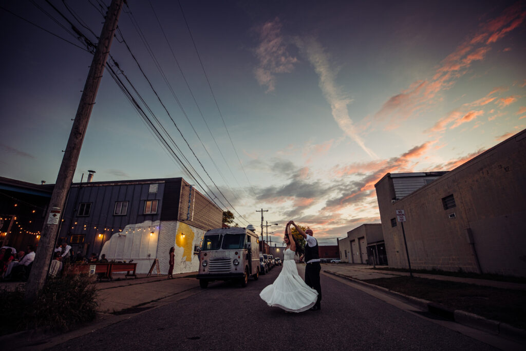 Bride and groom dancing in the street at sunset outside State Line Distillery in Madison with colorful evening sky.