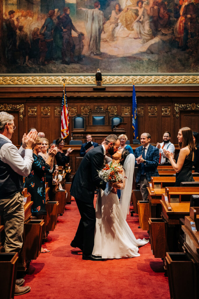 Bride and groom kissing in Wisconsin State Capitol chamber aisle as guests stand and applaud during intimate wedding ceremony.