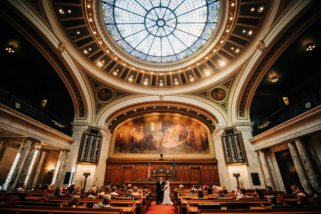 Wide view of wedding ceremony inside Wisconsin State Capitol rotunda with ornate dome and seated guests in Madison.