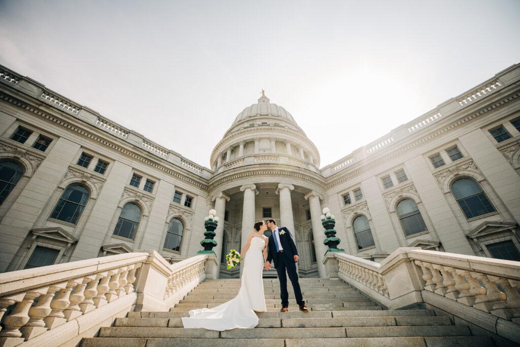 Bride and groom standing on steps outside Wisconsin State Capitol building in Madison with domed architecture behind them.