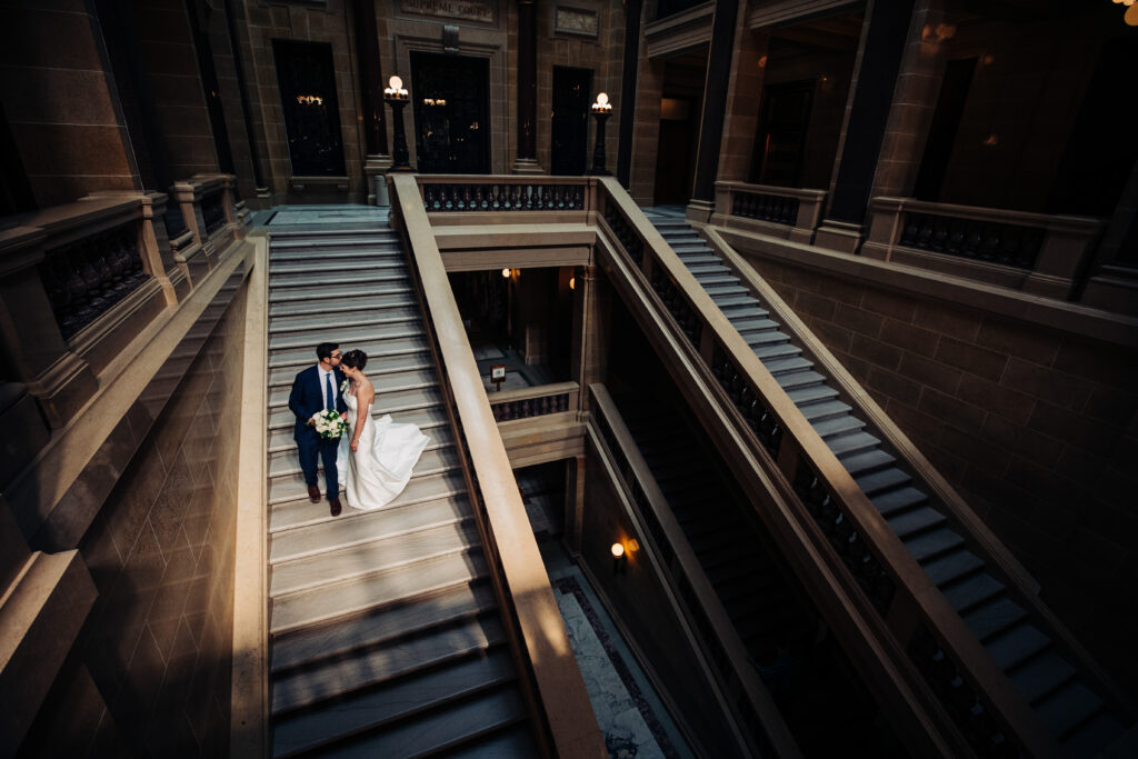 Bride and groom standing together on grand staircase inside Wisconsin State Capitol in Madison with dramatic architectural lines.