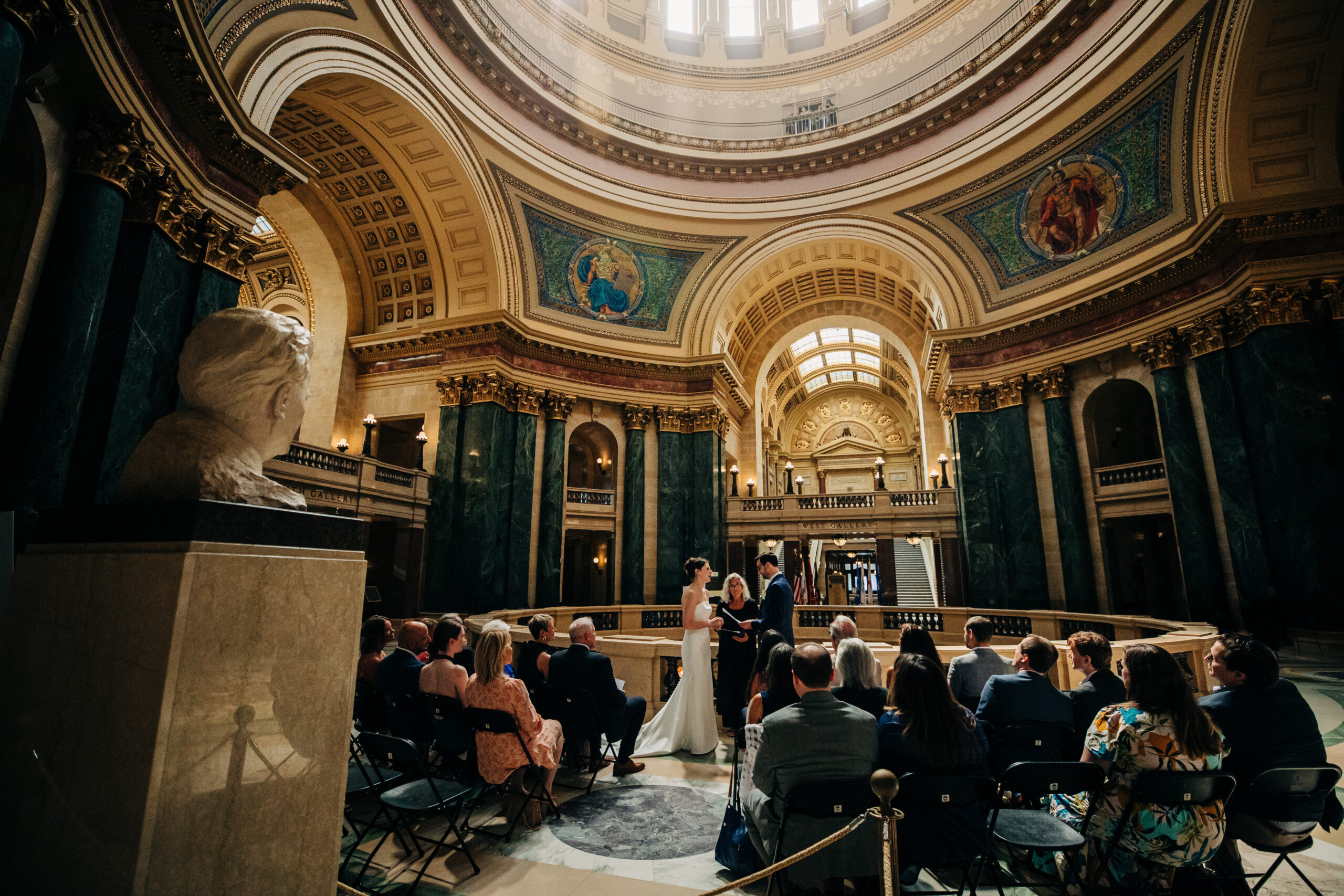 Couple exchanging vows during intimate wedding ceremony inside Wisconsin State Capitol rotunda in Madison with guests seated.