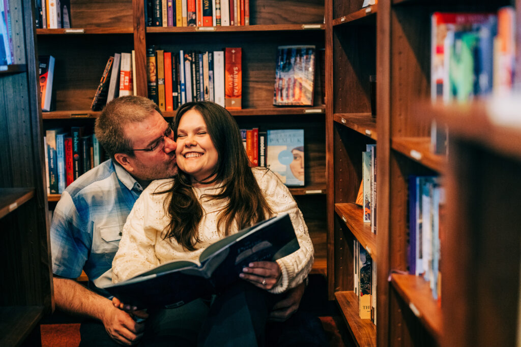 Couple sitting in front of bookshelves being affectionate and happily reading a book during their engagement photos in Madison, WI.