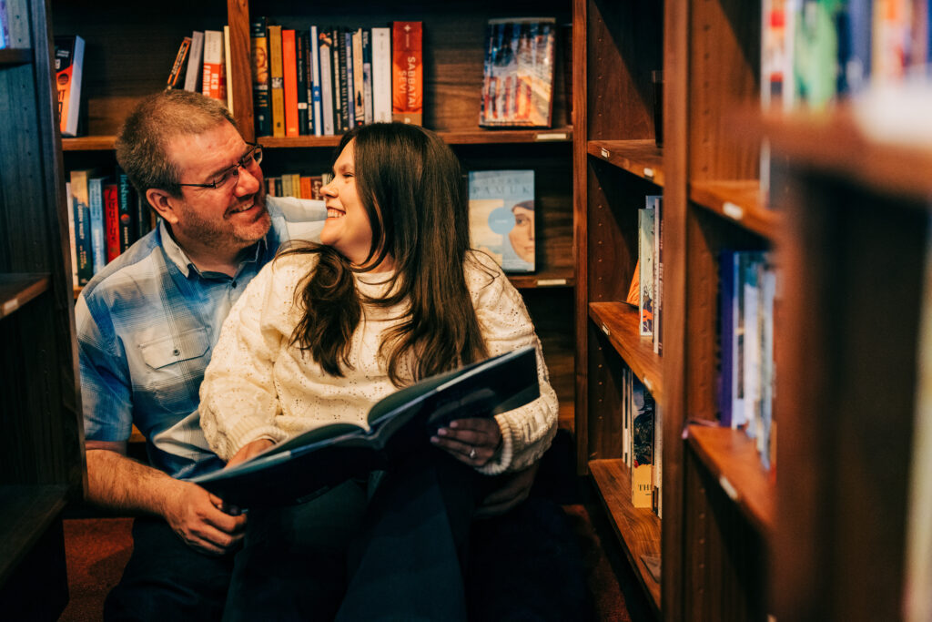 Couple sitting in front of bookshelves smiling and reading a book during their Leopolds Madison engagement photos in Madison, WI.