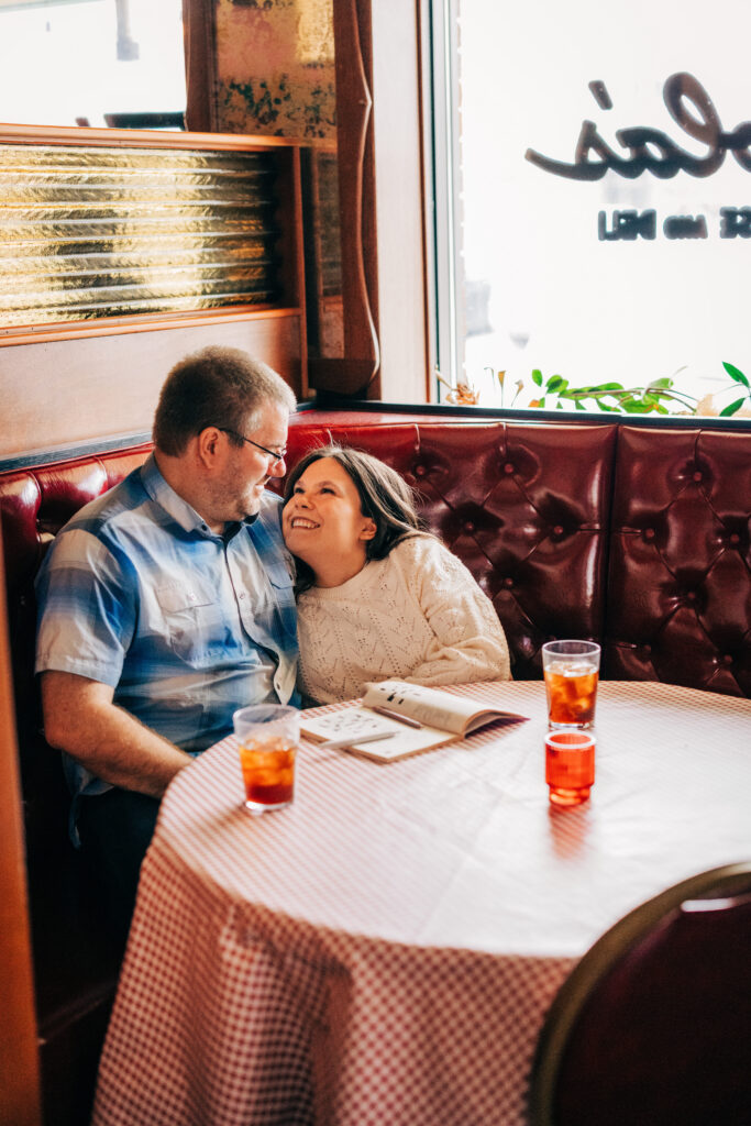 Couple in round booth snuggled and smiling with crossword puzzle at Leopold's Books Bar Caffè during Leopolds Madison engagement session in Madison, WI