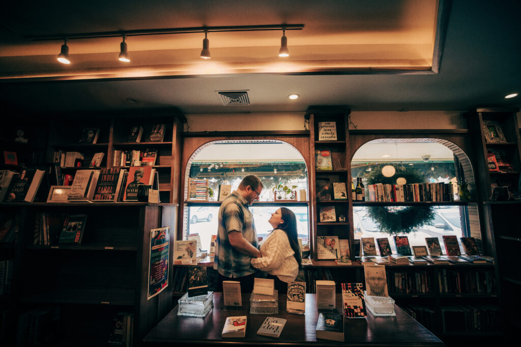 Couple facing each other holding hands among lots of books and two windows at Leopold's Books Bar Caffè during Leopolds Madison engagement session in Madison, WI