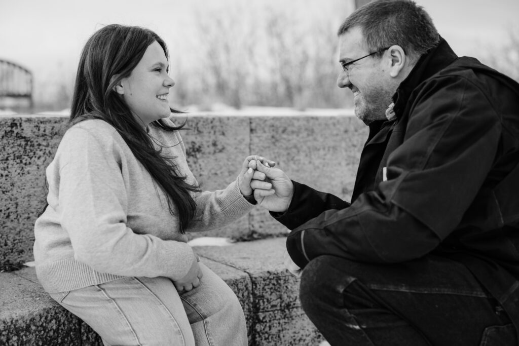 Man on one knee proposing in front of woman sitting on stone steps outside during engagement session at Picnic Point
