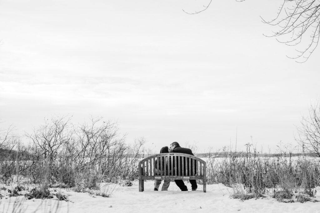 Couple kissing and sitting on bench in front of frozen lake during engagement session at Picnic Point