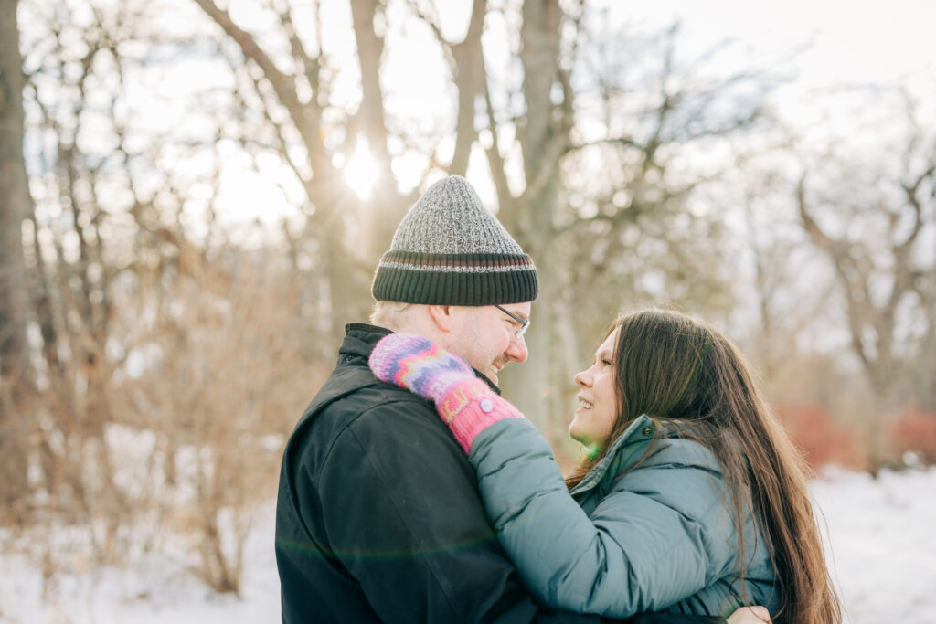 Couple facing each other smiling by trees in the snow during engagement session at Picnic Point