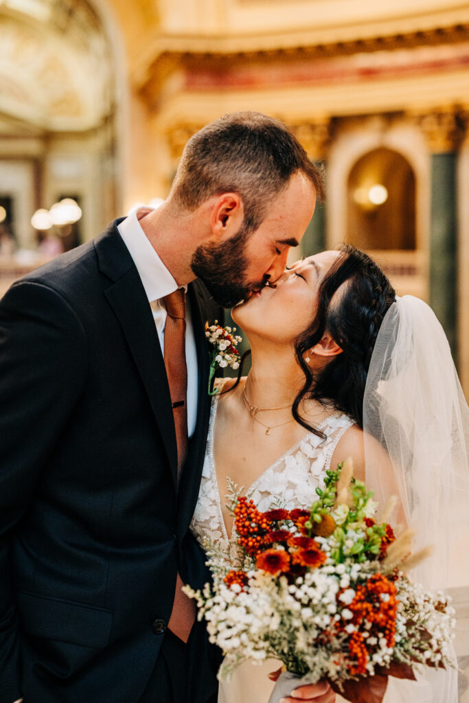 Bride and groom kissing while holding an autumn-inspired wedding bouquet inside the Wisconsin State Capitol.