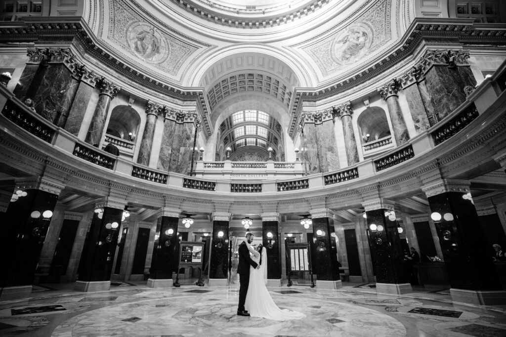 Wide black-and-white portrait of newlyweds standing beneath the grand Wisconsin State Capitol rotunda.