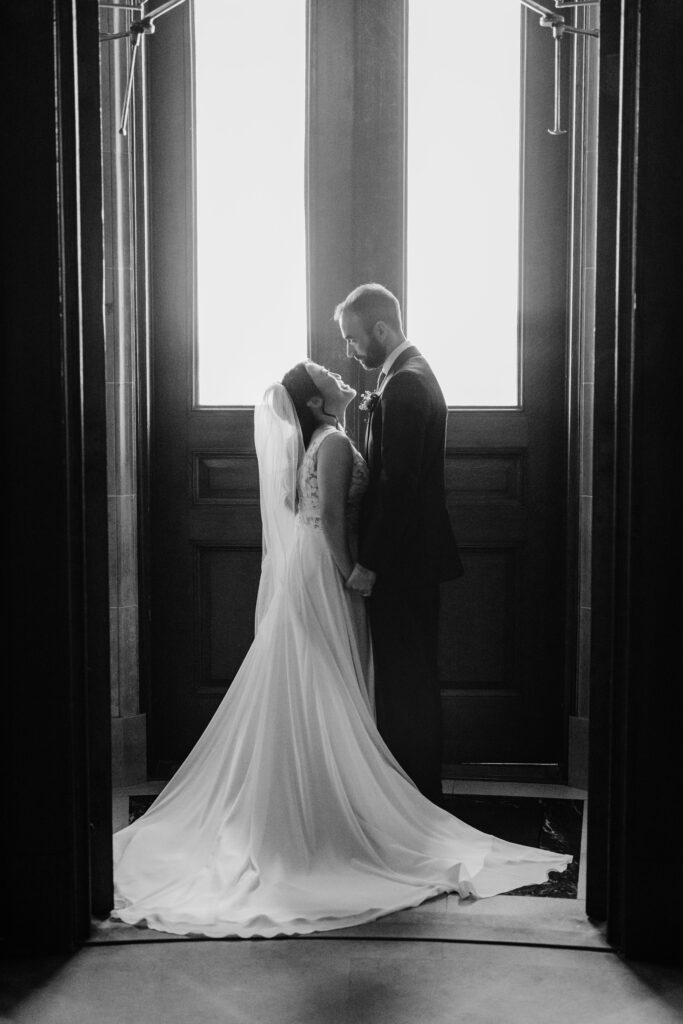 Black-and-white portrait of bride and groom standing hand in hand beneath tall Capitol windows during Wisconsin wedding.