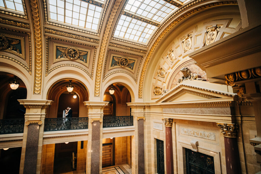 Bride and groom kissing beneath ornate arches and chandeliers inside the Wisconsin State Capitol.