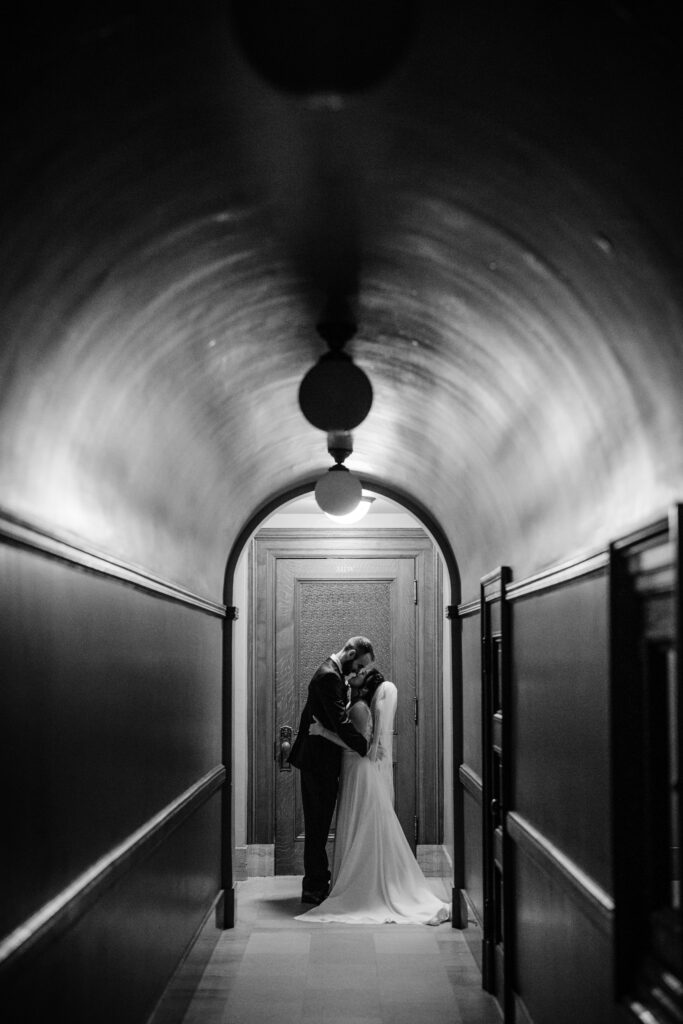 Black-and-white wedding portrait of couple embracing in dimly lit Capitol hallway.