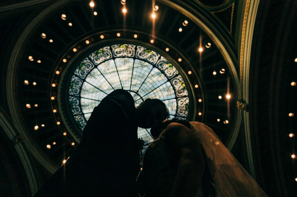 Romantic silhouette of bride and groom kissing beneath stained-glass Capitol dome skylight.