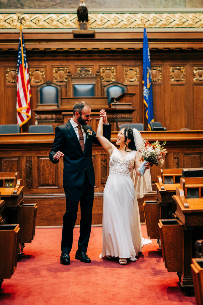 Newly married couple celebrating inside Wisconsin State Capitol courtroom with bouquet and raised hands.