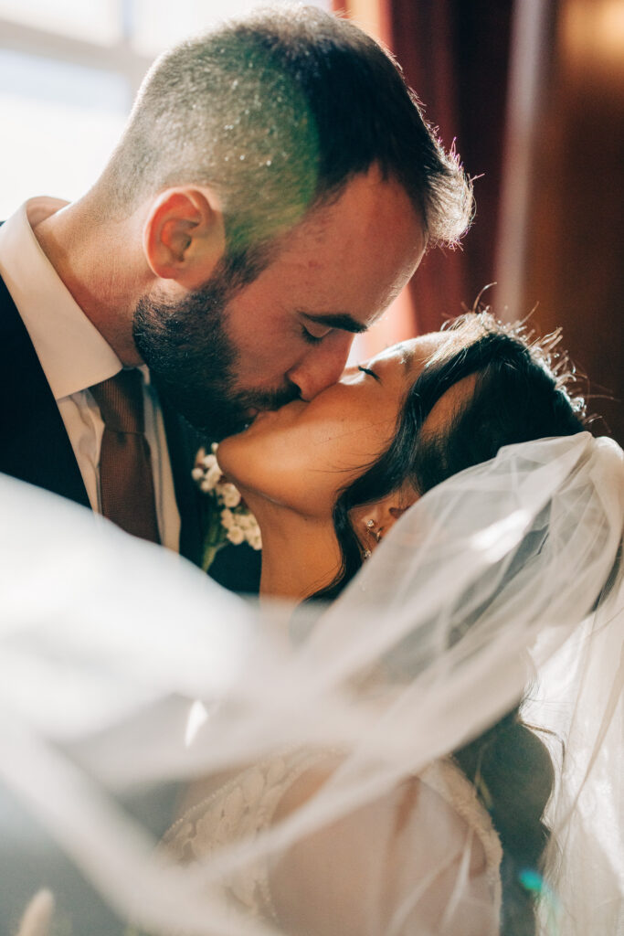 Close-up wedding kiss portrait with soft veil movement and warm natural light.