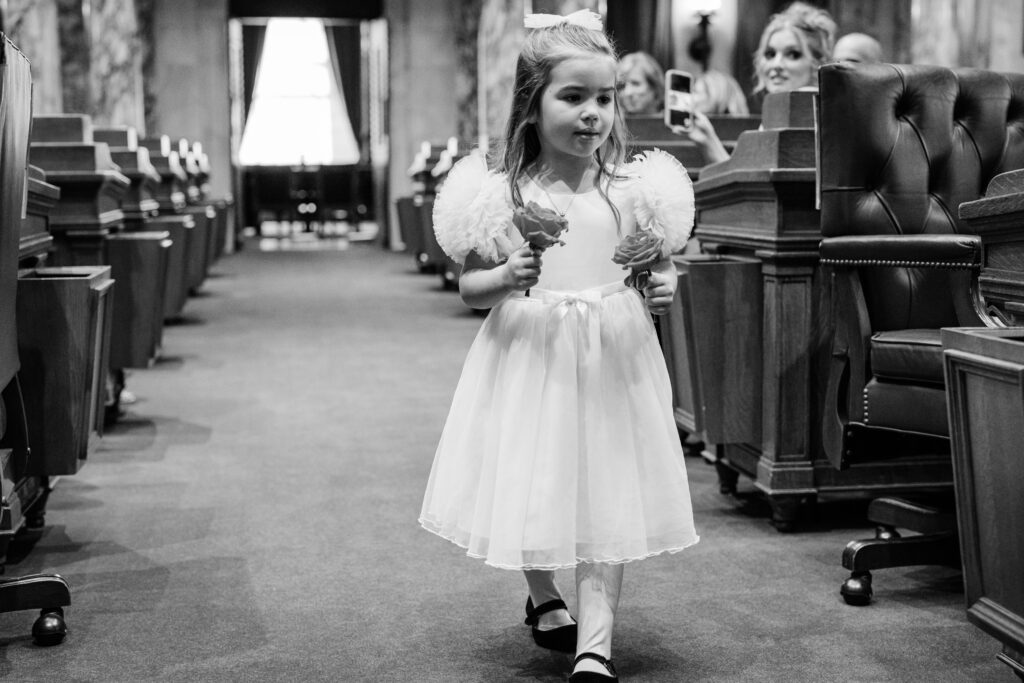 Flower girl walking down the aisle in a white dress holding roses during an intimate courthouse wedding ceremony.