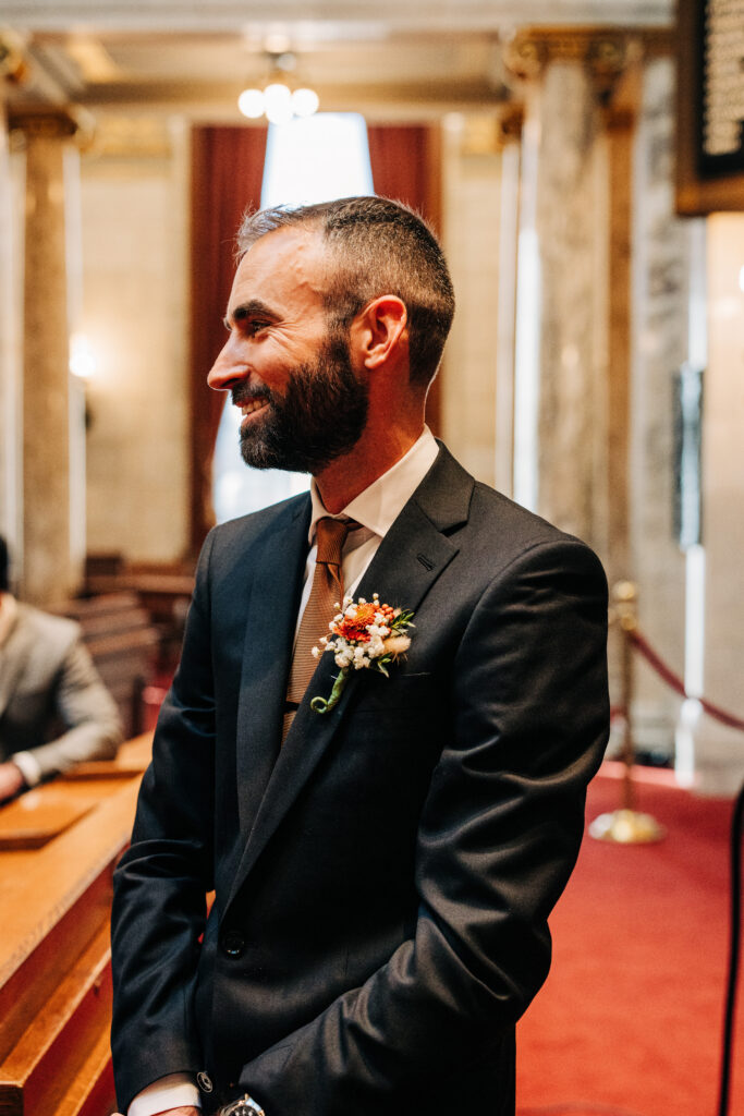 Groom in a dark suit and brown tie smiling during a courthouse wedding ceremony with marble columns behind him.