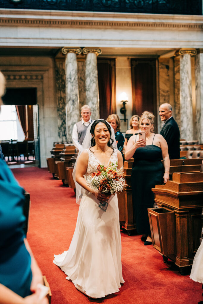 Bride walking down the aisle holding a fall bouquet inside an ornate courthouse wedding venue with guests watching.