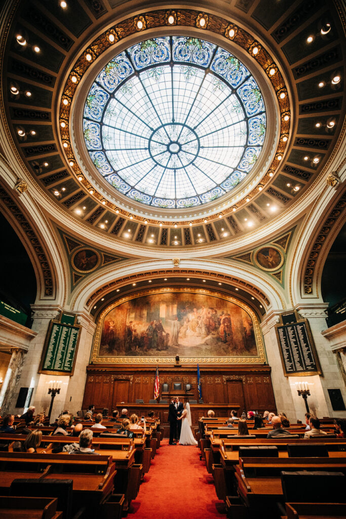Wide view of a courthouse wedding ceremony beneath an ornate stained glass dome in a historic capitol building.