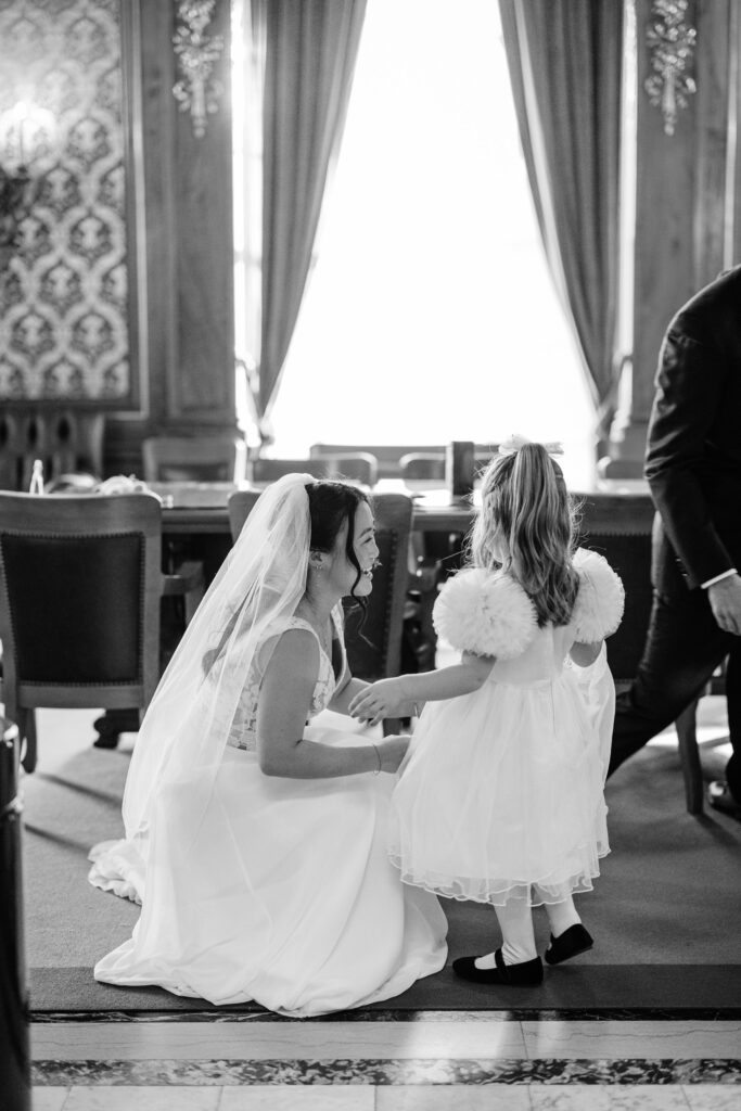 Bride kneeling beside a flower girl in a white dress inside a softly lit courthouse wedding venue.