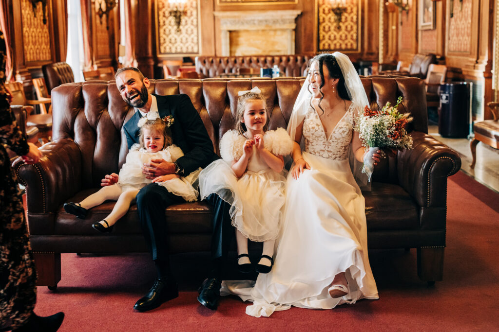 Bride and groom seated on a leather couch with two flower girls after their intimate courthouse wedding ceremony.