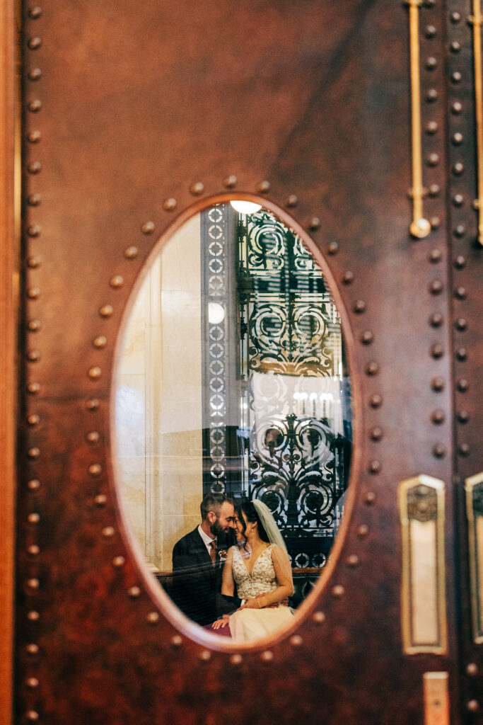 Bride and groom reflected through an oval window in a historic wooden courthouse door during wedding portraits.