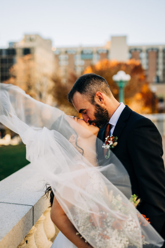 Bride and groom kissing outdoors with flowing veil and autumn colors during romantic courthouse wedding portraits.