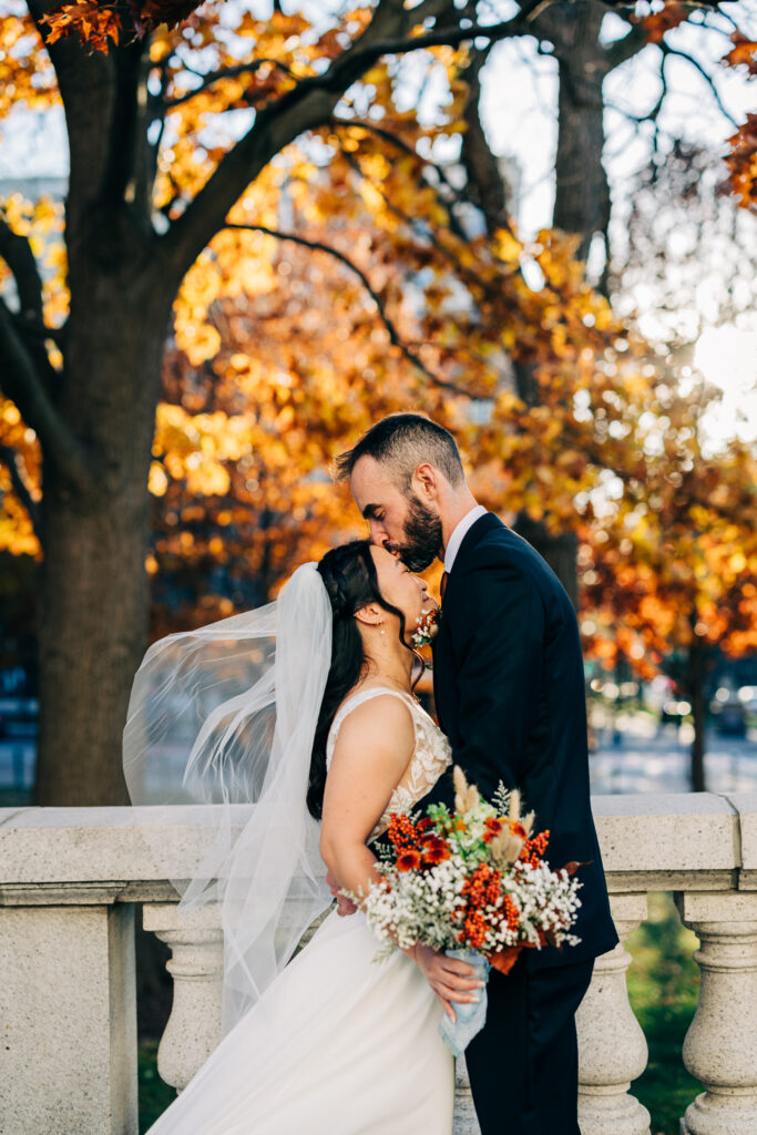 Bride and groom sharing a kiss outdoors with autumn trees and flowing veil during fall wedding portraits.