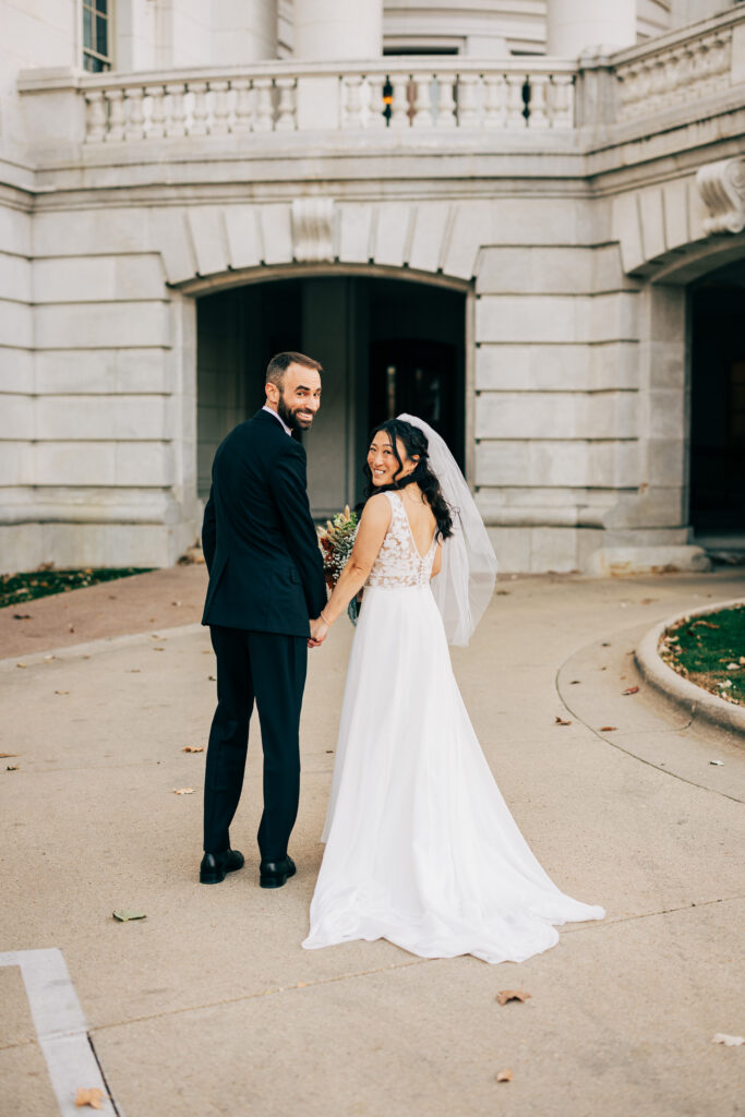 Bride and groom holding hands and looking back while walking outside a historic courthouse after their ceremony.