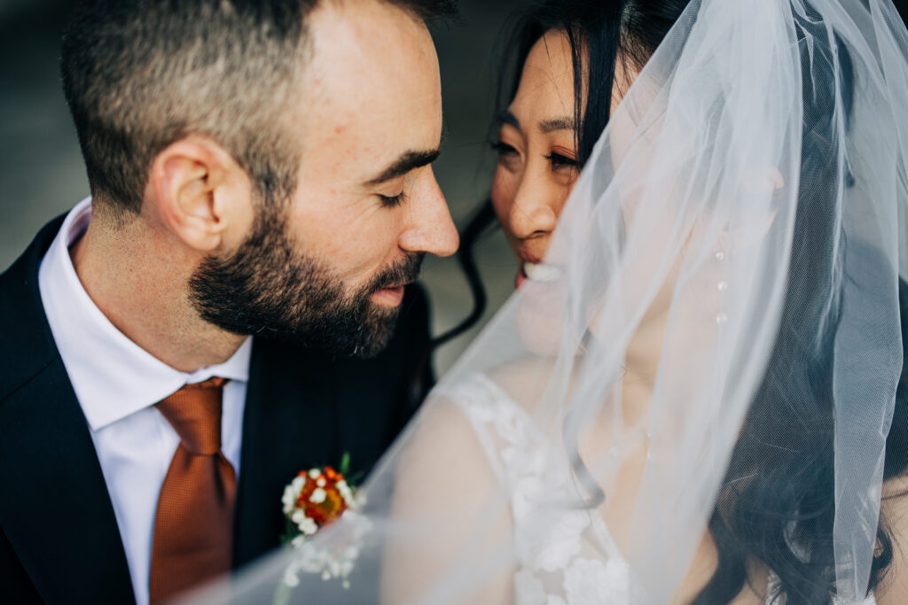 Bride and groom smiling closely beneath a sheer veil during romantic courthouse wedding portraits outdoors.