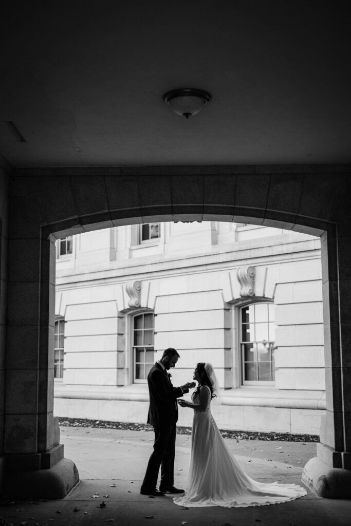 Silhouette of bride and groom standing beneath a stone archway outside a historic courthouse after their wedding.