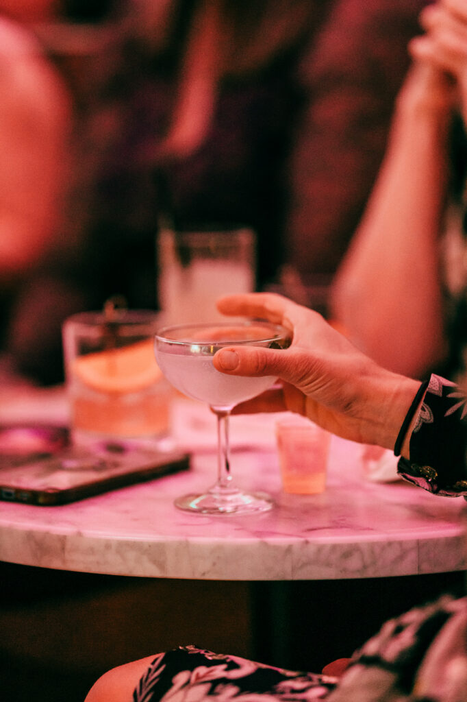 Close-up of a hand holding a coupe cocktail glass on a marble table inside a dimly lit lounge.