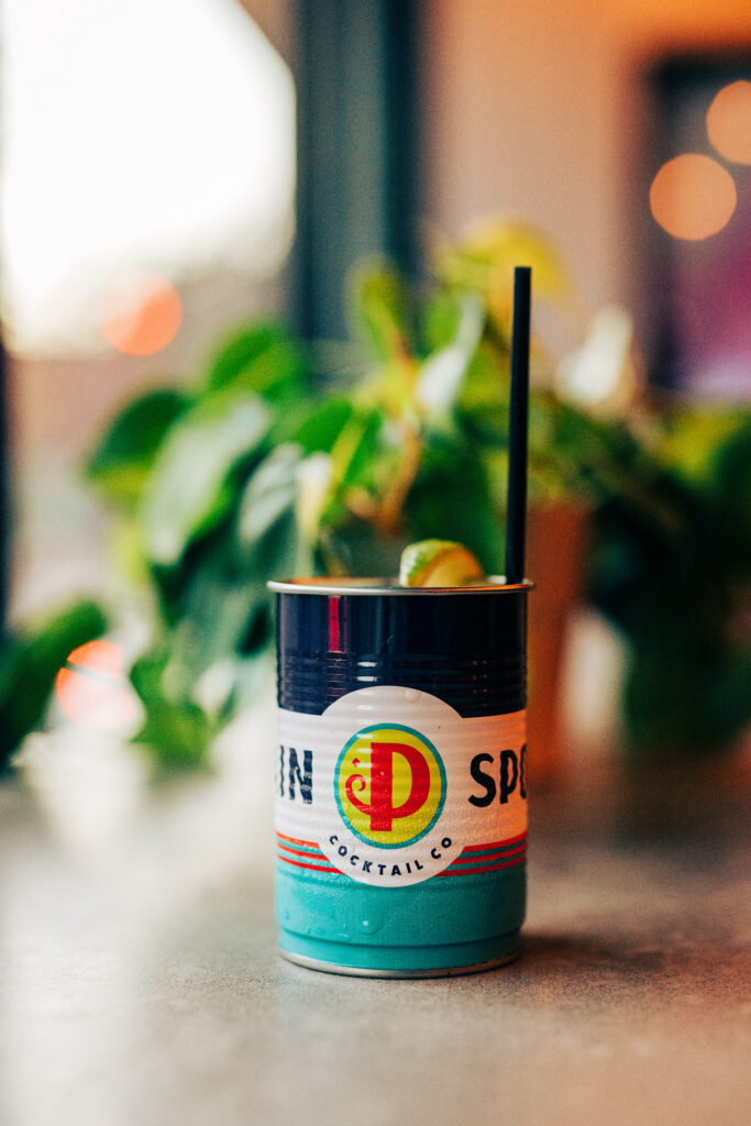 Canned cocktail with lime garnish and straw on a bar counter with soft neon lighting and plants in the background.