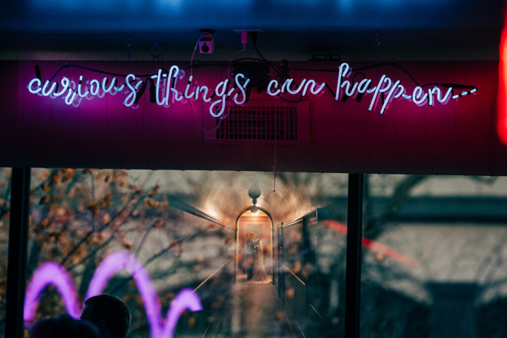 Bride and groom reflected in a neon-lit cocktail bar window beneath a glowing “curious things can happen” sign.