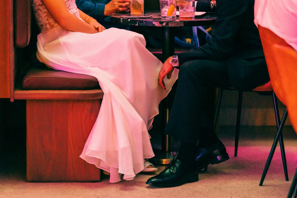 Bride and groom seated in a modern cocktail lounge with pink neon lighting and drinks on a small table.
