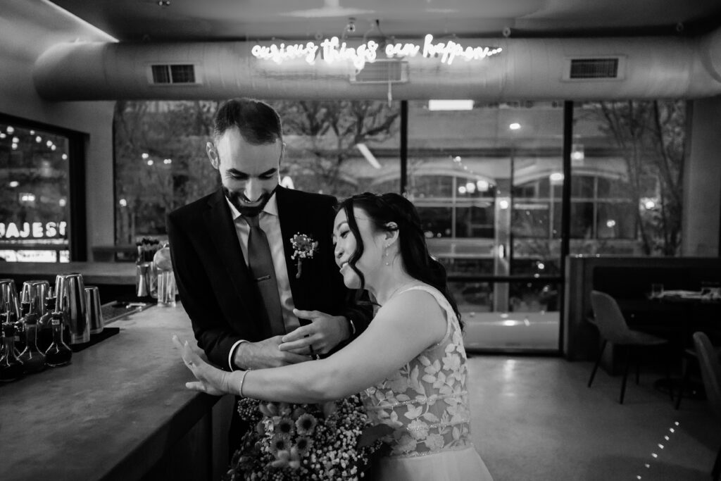 Black-and-white portrait of bride and groom leaning against a cocktail bar beneath a glowing neon sign.