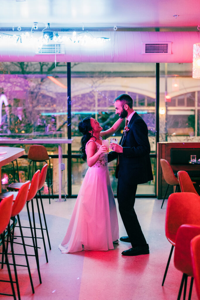 Bride and groom dancing with cocktails in hand inside a colorful neon-lit lounge after their wedding.